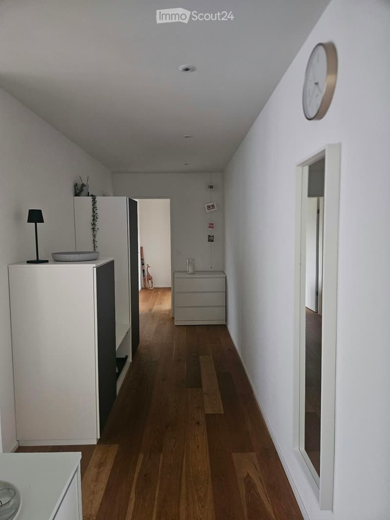 Modern hallway with wooden flooring, white walls, and minimal decor including a small lamp, a bowl, and a clock on the wall.