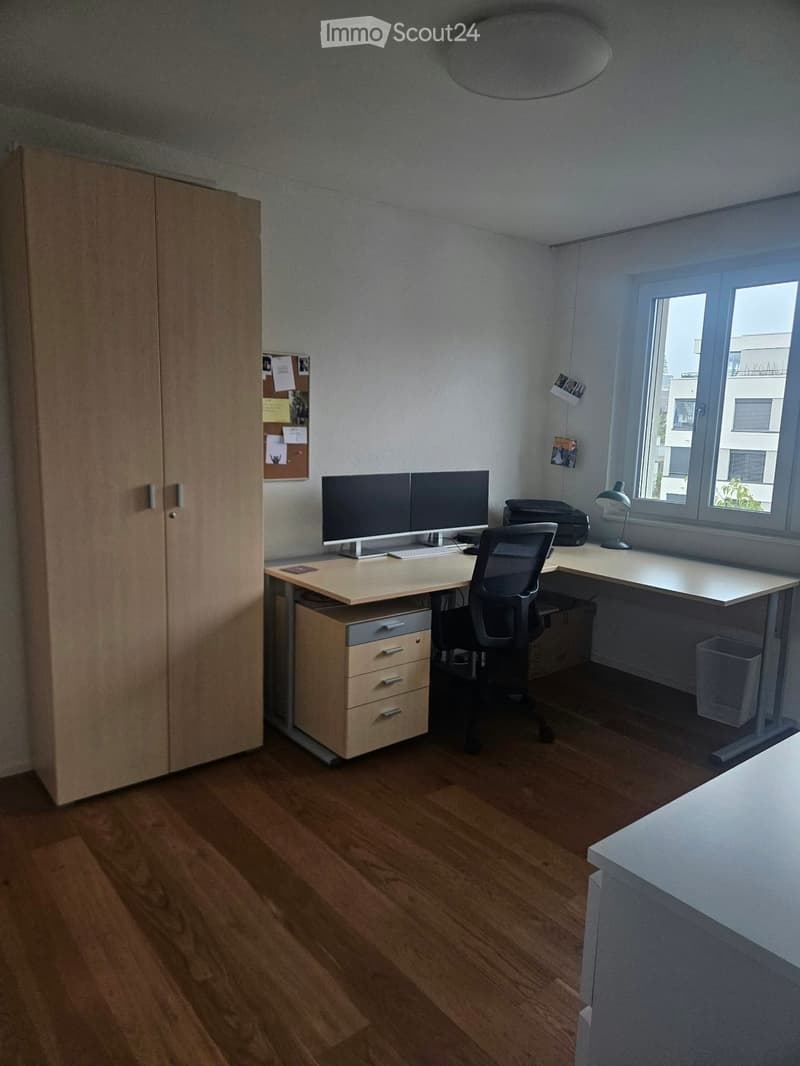 A modern, well-lit office space with a large wooden desk, ergonomic chair, dual monitors, printer, and a tall cabinet. The room features light wooden flooring and large windows with a view of the city.