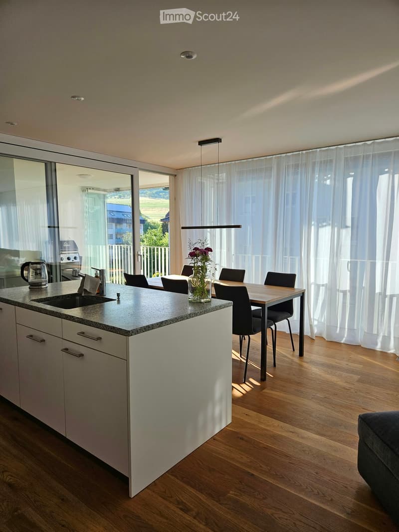 Modern kitchen with a white island, granite countertop, sink, coffee maker, and a dining area with a wooden table and chairs. Large windows with sheer curtains provide natural light.
