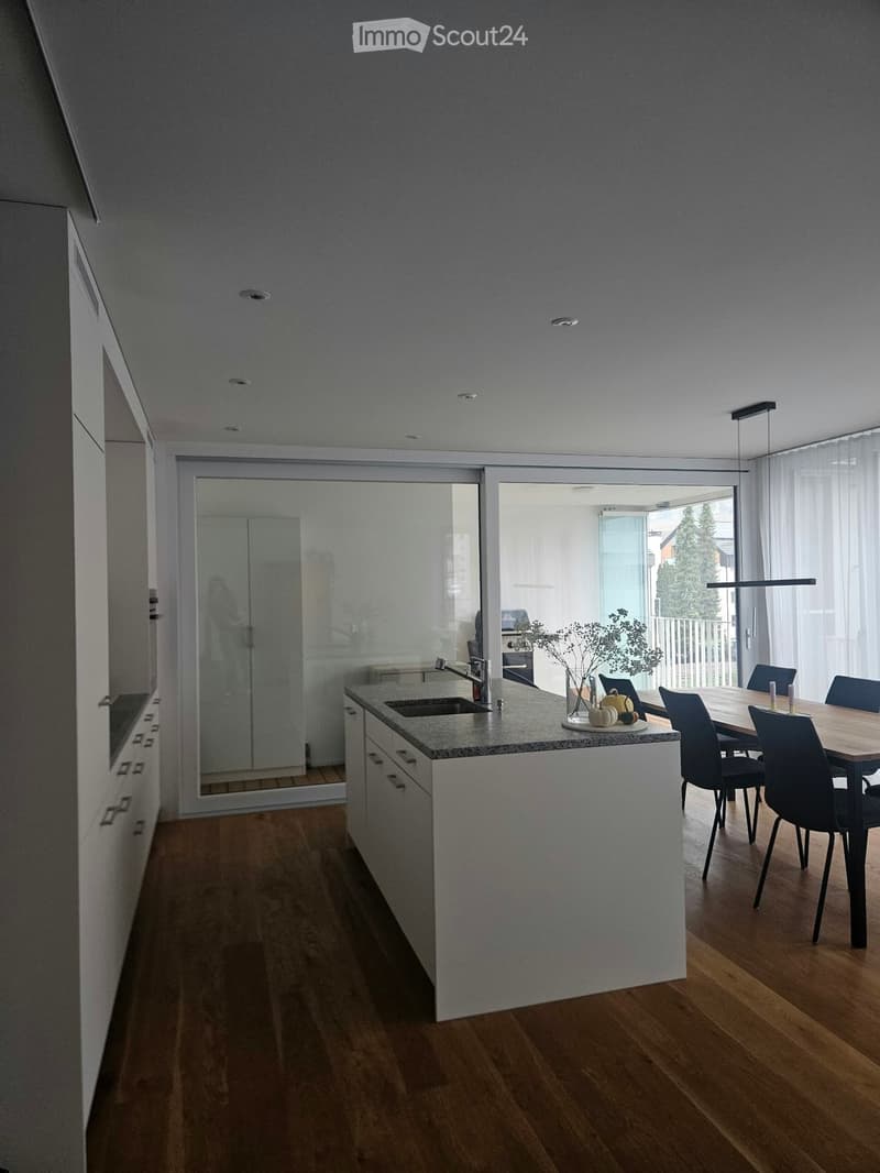 Modern kitchen with a central island, sink, and a dining area with a table and chairs. The space features wooden flooring, white cabinets, and large windows with sheer curtains.