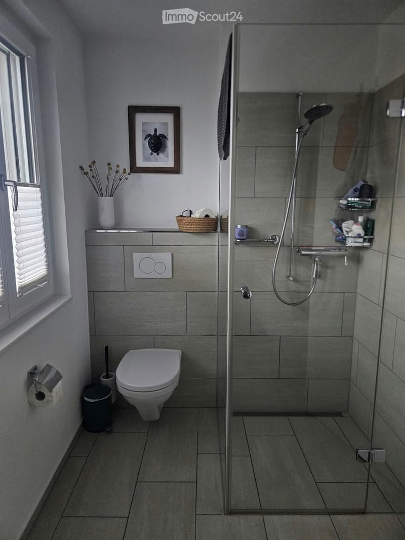 Modern bathroom with grey tiles, a toilet, a glass-enclosed shower, a framed picture, a vase with dried flowers, and a wicker basket on the counter.