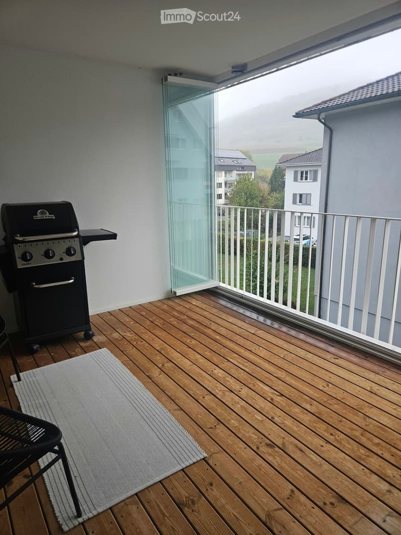 Balcony with wooden decking, glass sliding door, white railing, grill, chair, and rug.