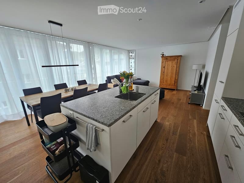 Open-plan kitchen and living area with white cabinets, granite countertop, wooden dining table, sofa, and large windows with white curtains.