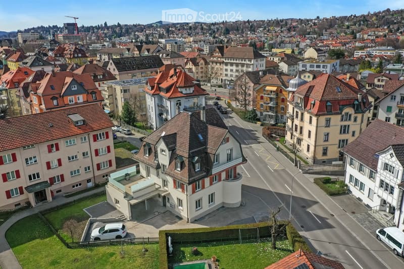 Aerial view of a residential neighborhood with multiple buildings, red-tiled roofs, and a mix of modern and traditional architecture. The area includes streets, parked cars, and a small garden with a swimming pool.