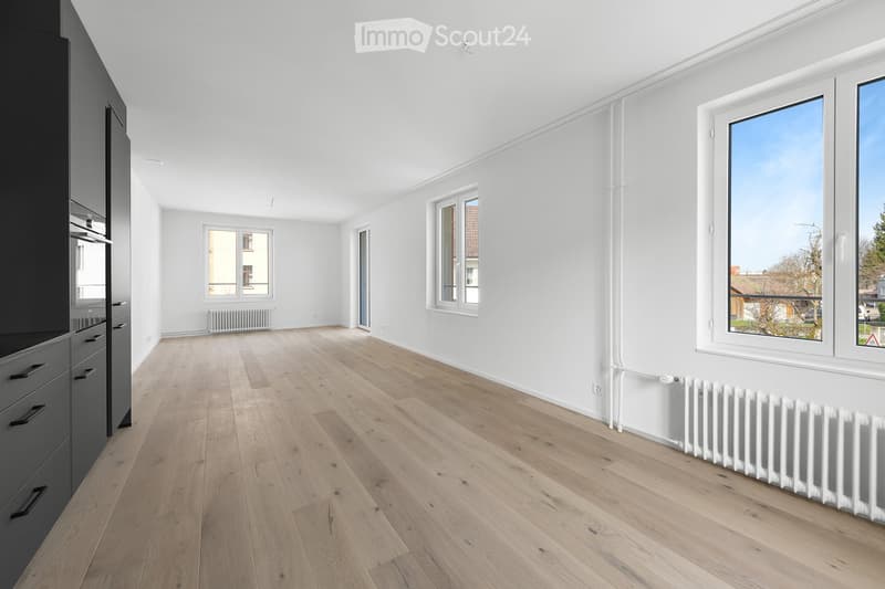Empty room with light wooden flooring, white walls, multiple windows, and radiators. No furniture or furnishings present.