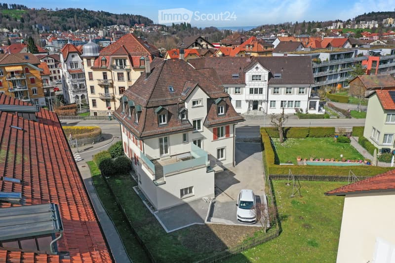Aerial view of a residential neighborhood with multiple houses, red-tiled roofs, and a mix of architectural styles. A white car is parked in a driveway, and there is a small garden area with a swimming pool and potted plants.