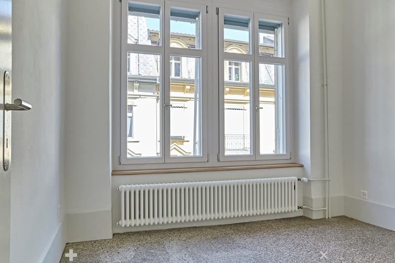 Empty room with white walls, carpeted floor, large windows with wooden sills, radiator, and a door handle visible on the left side.