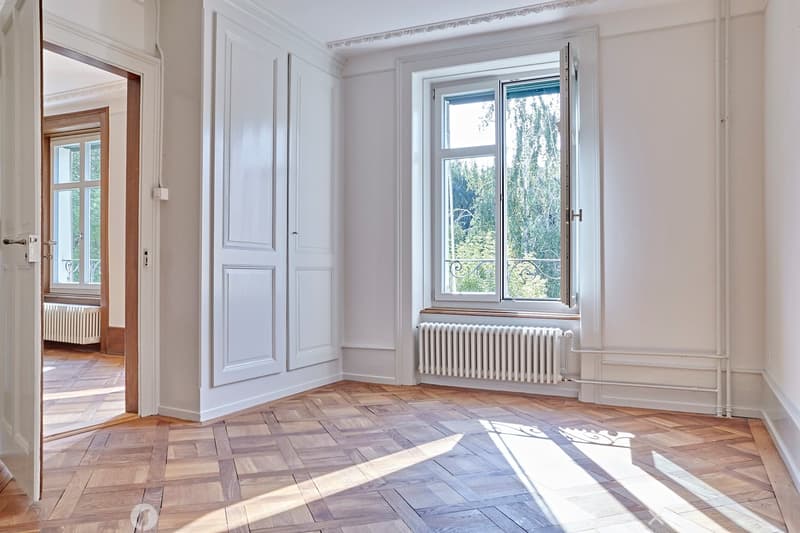 Empty room with light-colored walls, wooden flooring, and large windows allowing natural light to enter. The room features radiators and a door leading to another space.