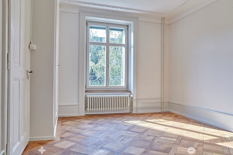 Empty room with wooden herringbone flooring, white walls, large window with double glass panels, radiator below the window, and a door on the left side.