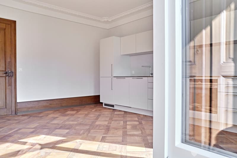 Modern kitchen with white cabinets, wooden herringbone floor, and a sleek faucet. The space is bright and minimalist, with a wooden door visible on the left.