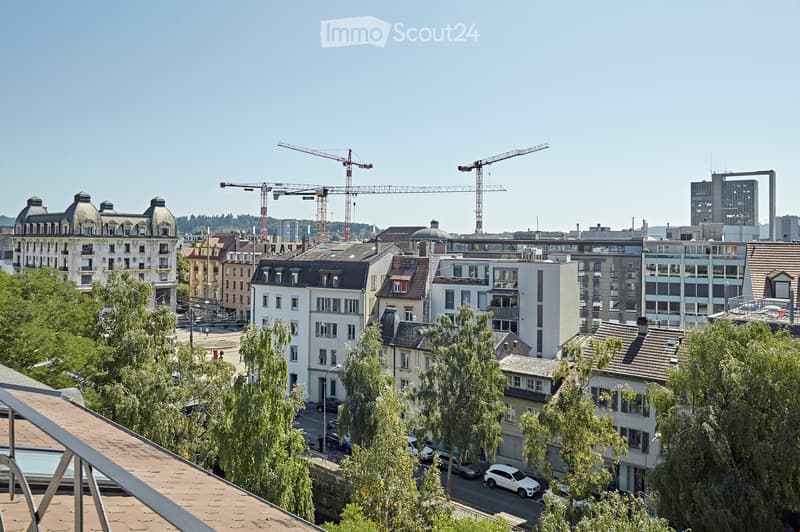 A panoramic view of a cityscape with a mix of residential and commercial buildings, including multi-story structures with varied architectural styles, surrounded by trees and visible construction cranes in the background.