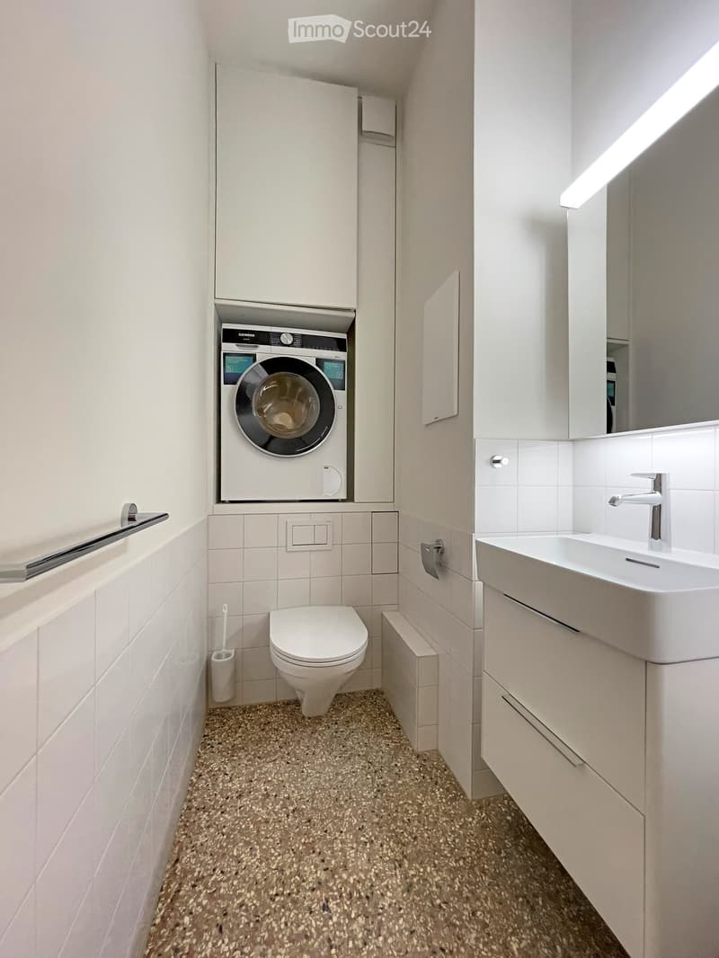 Modern bathroom with white tiles, a toilet, a sink with a modern faucet, a built-in washing machine, and a handrail for accessibility.