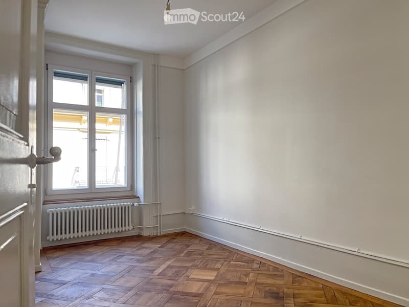 Empty room with wooden herringbone flooring, white walls, large window with double panes, radiator, and a single light fixture on the ceiling.