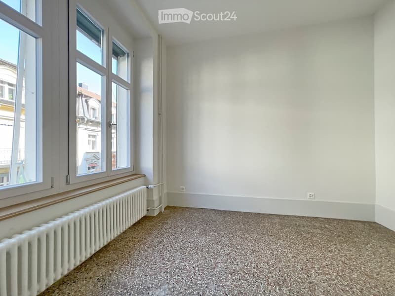 Empty room with white walls, carpeted floor, radiator, and large windows with wooden sills.
