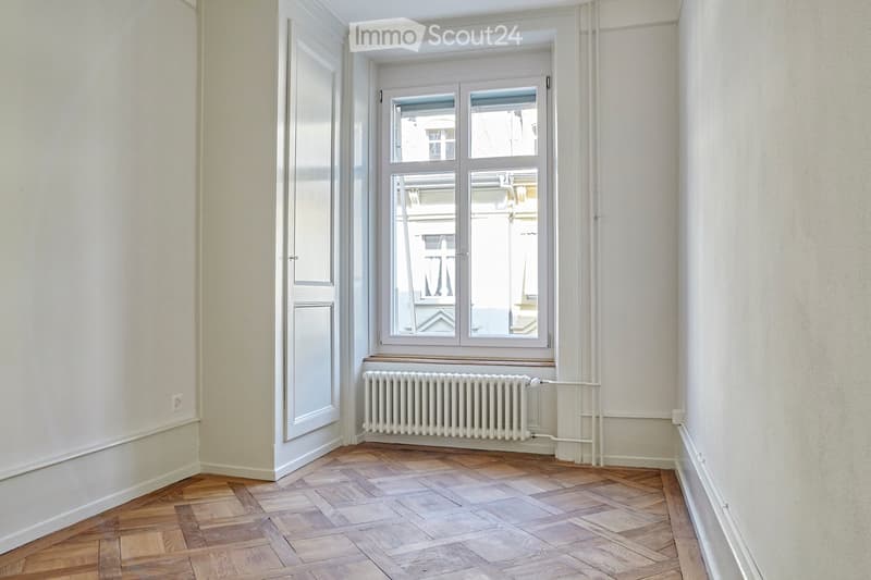 Empty room with wooden herringbone flooring, white walls, a window with white frame, and a radiator below the window.