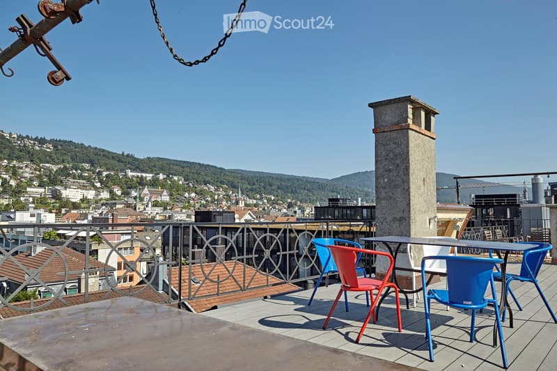 A rooftop terrace with a metal railing, overlooking a cityscape and hills. The terrace features a small table and colorful chairs.