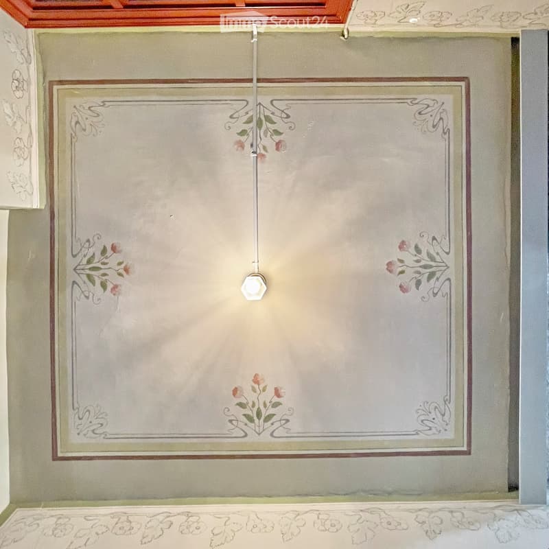 An unfurnished room with light-colored walls and decorative floral patterns, featuring a single light fixture hanging from the ceiling.