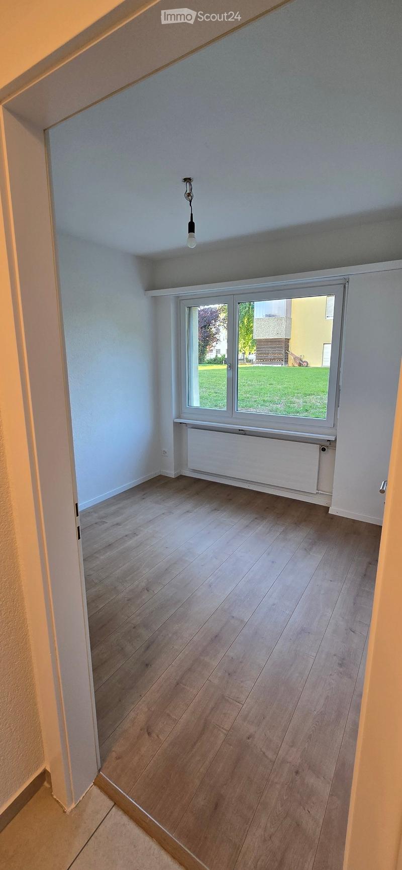 Empty room with wooden flooring, white walls, large window with view of green area, radiator, and exposed ceiling beams.