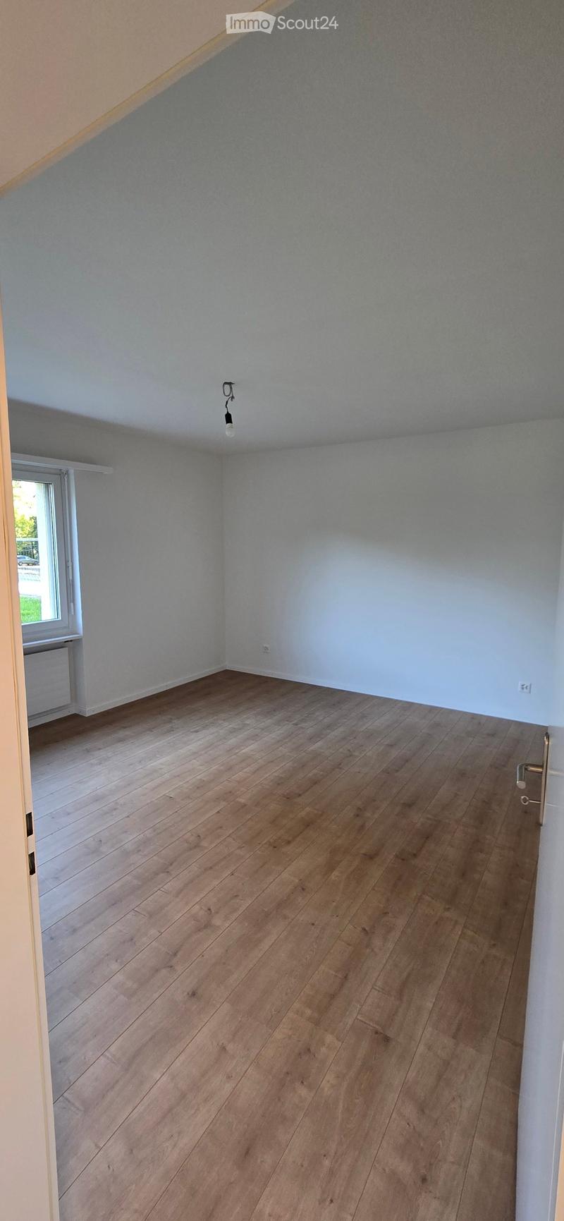 Empty room with wooden flooring, white walls, and a single window with a radiator below it. The room has a simple, clean design with no furniture or decorations.