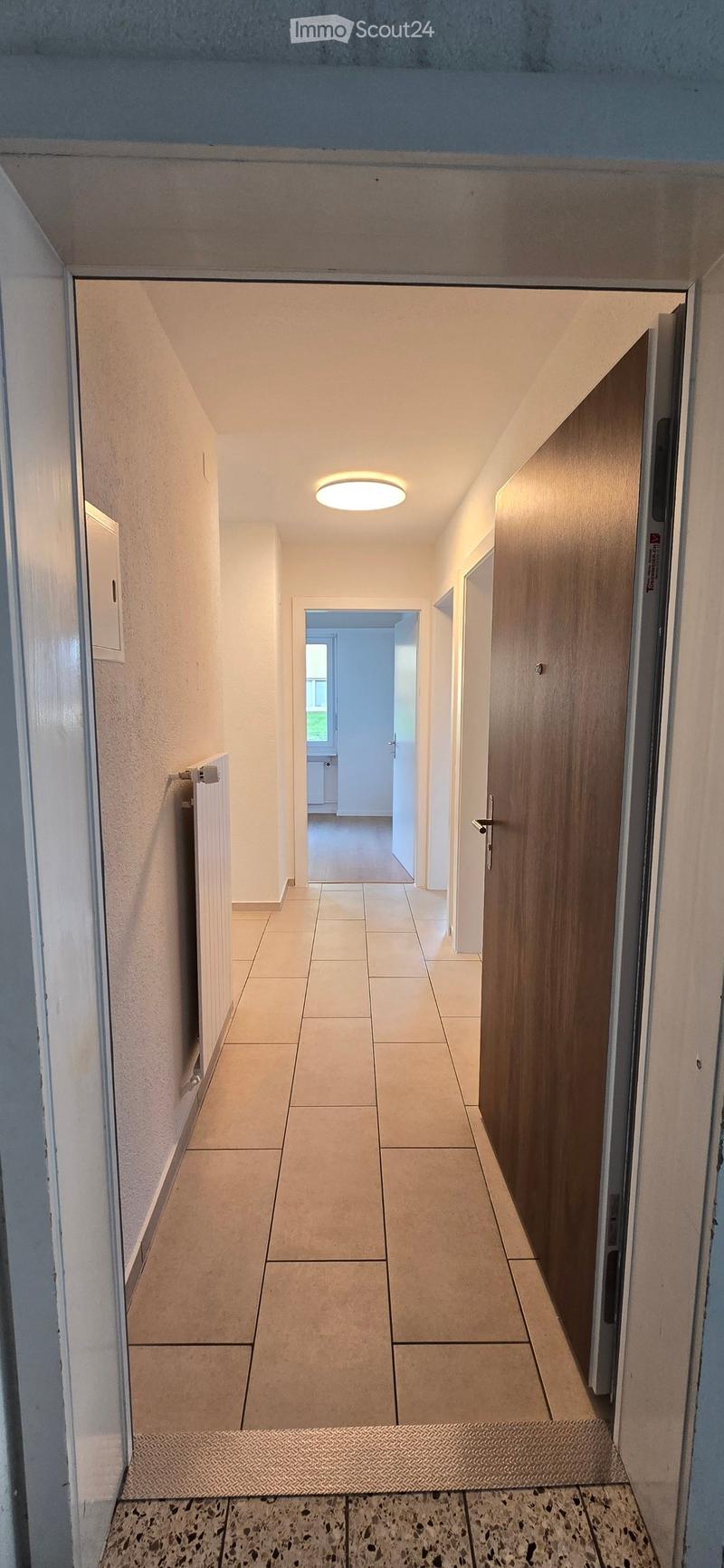 Clean, well-lit hallway with tiled flooring, white walls, and a recessed ceiling light. Features a wooden door on the right and a radiator on the left, leading to another room with visible furniture.