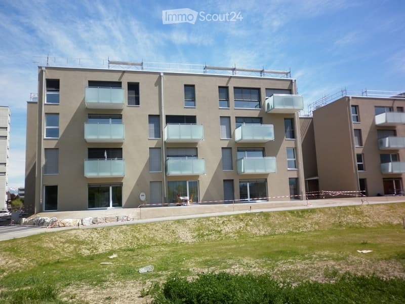 Modern multi-story residential building with balconies, large windows, and a raised ground floor. The building is partially under construction with visible scaffolding and a grassy area in front.