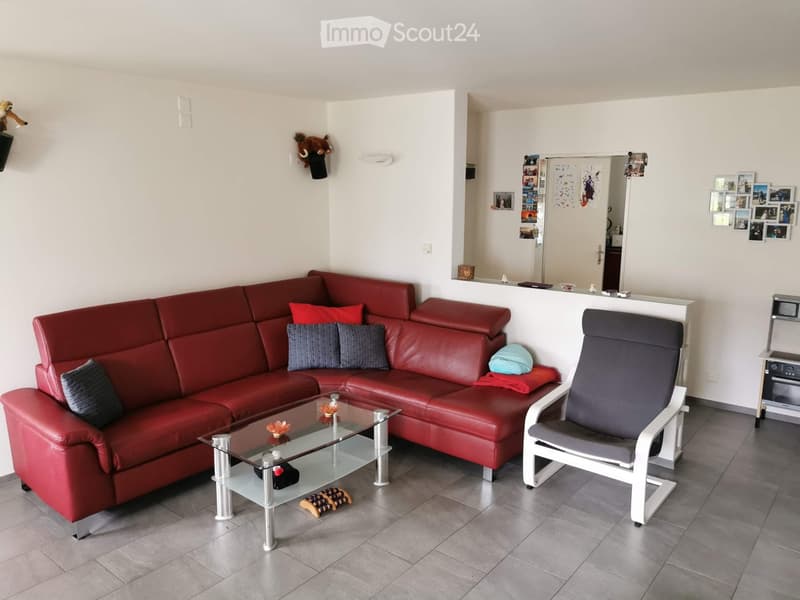 Modern living room with a red leather sectional sofa, glass coffee table, grey armchair, and a small table with a microwave. The room features grey tiled flooring and white walls, with decorative items and photos displayed on the walls.