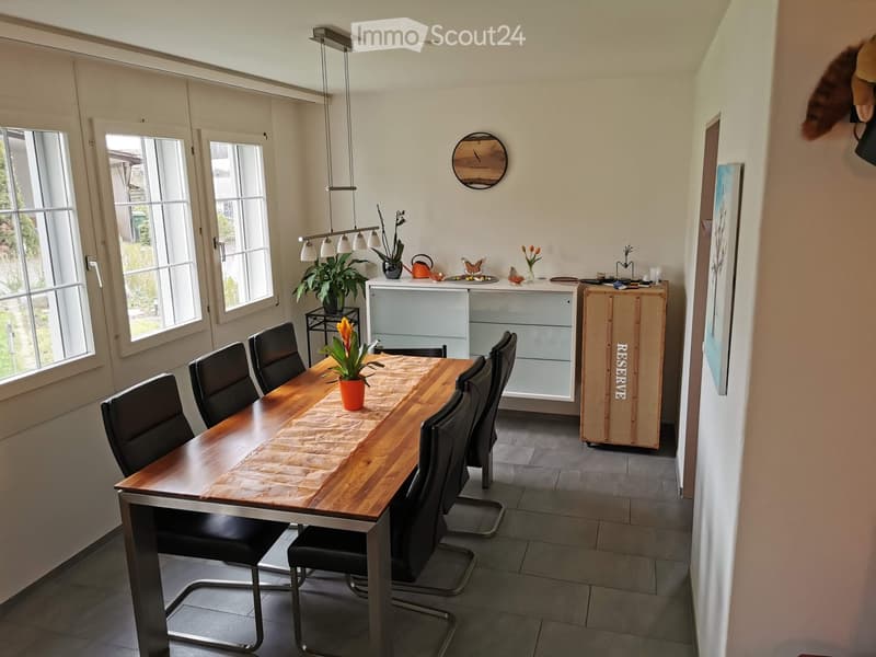Bright kitchen with a wooden dining table surrounded by black chairs, a potted plant centerpiece, and a modern cabinet with glass doors. Large windows provide natural light.