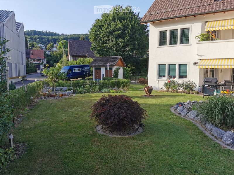 Well-maintained garden with a lawn, shrubs, trees, and a small decorative water feature. A modern house with a covered patio and outdoor furniture is visible in the background.
