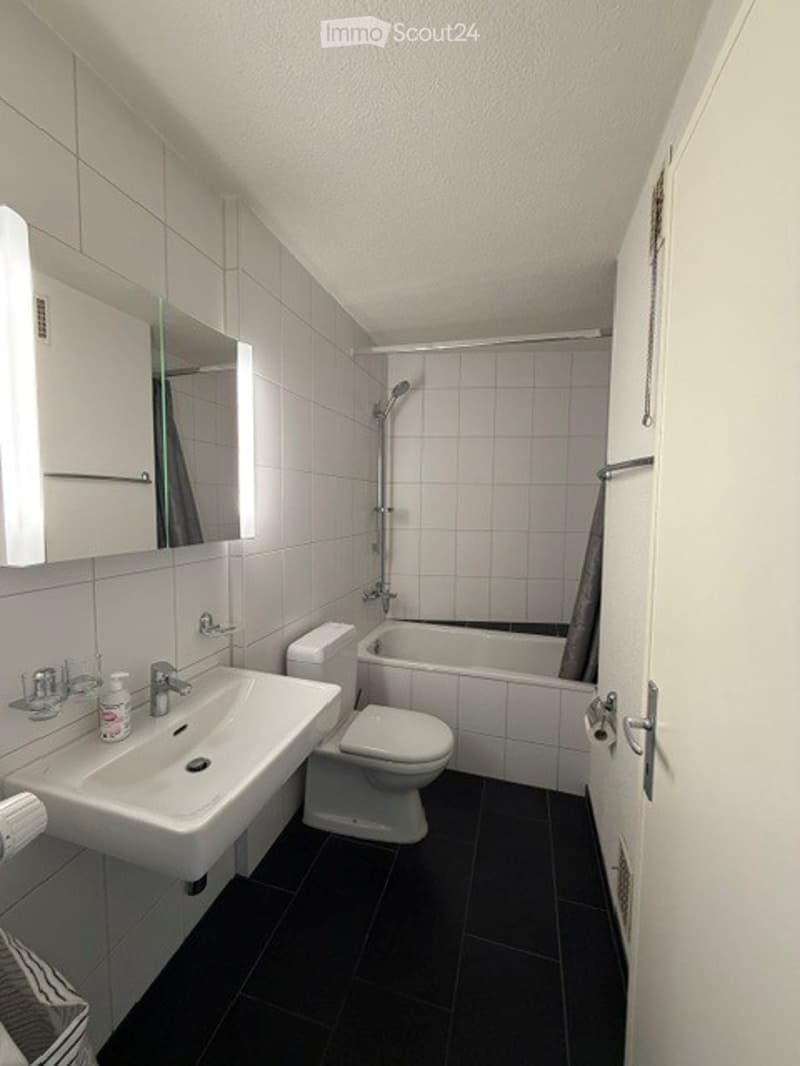 Modern bathroom with white tiles, black floor, sink, toilet, bathtub, and shower. Features a mirror and cabinet above the sink.