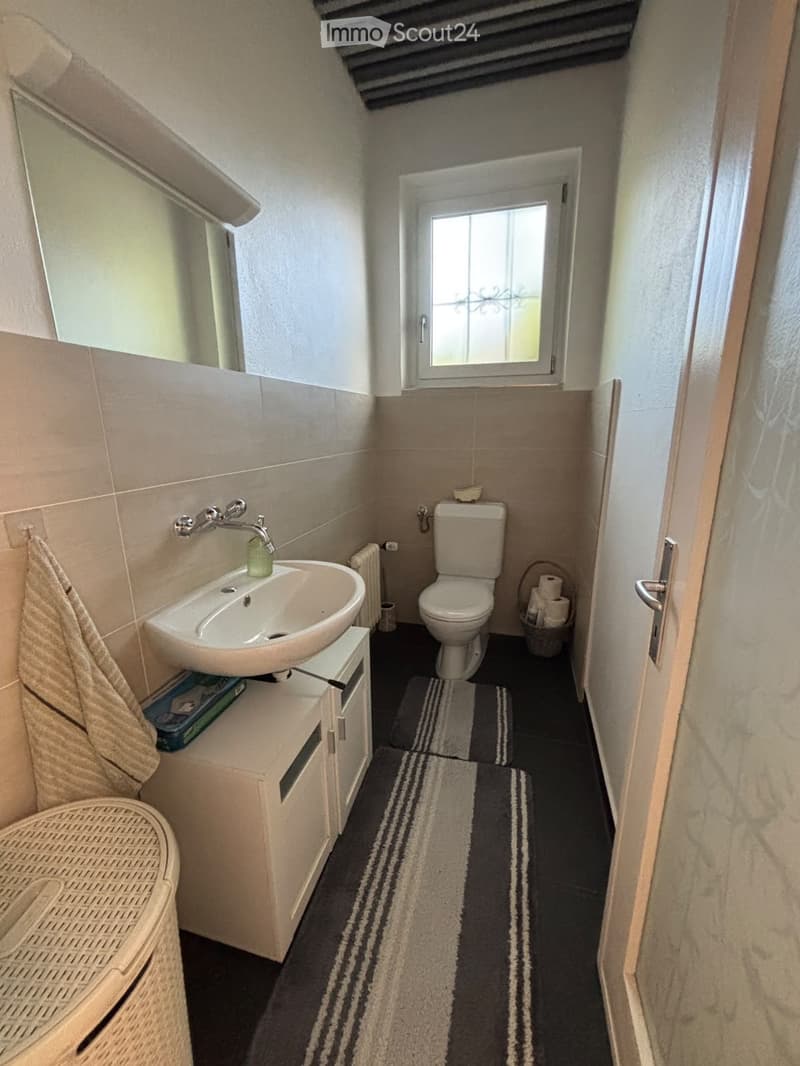 Cozy bathroom with a sink, toilet, mirror, and a wicker chair near the door. The room features a window with decorative glass and a striped rug on the floor.