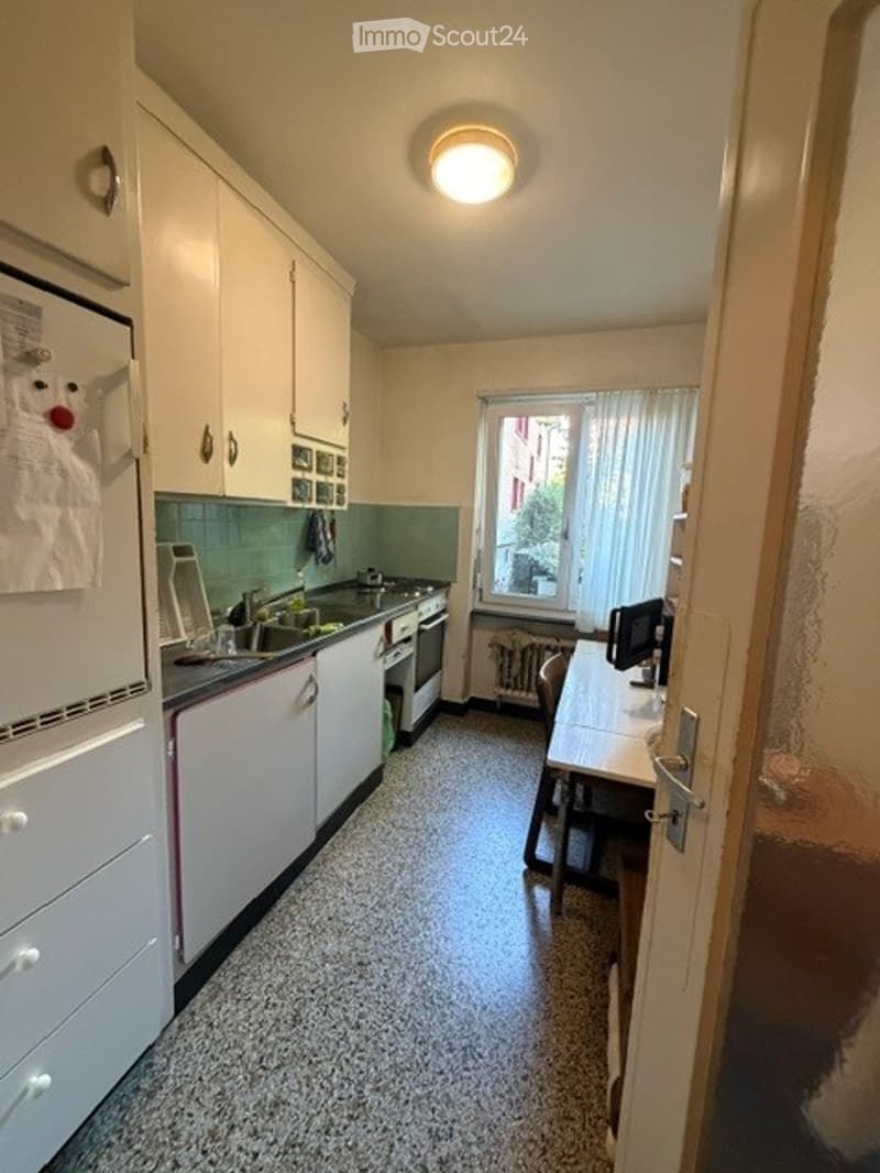 A compact kitchen with white cabinets, a refrigerator, sink, oven, and a small table with a chair and computer monitor. The space features a tiled floor and a window with curtains.