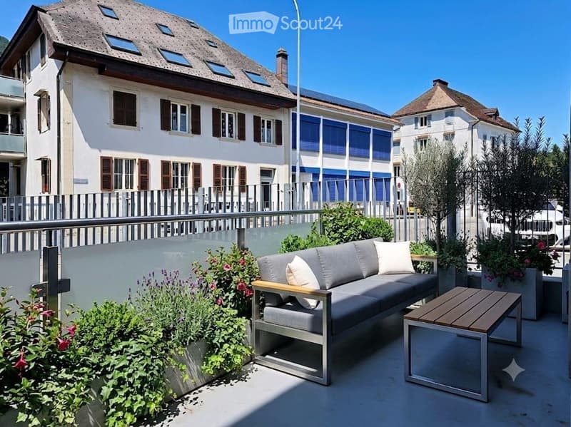 Balcony with a grey sofa, wooden table, and potted plants, overlooking a multi-story building with brown shutters and a metal fence.