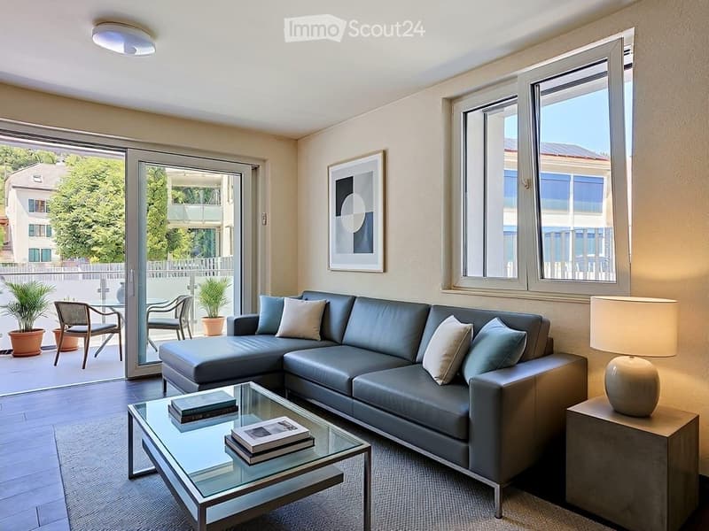 Modern living room with a grey sectional sofa, glass coffee table, side table with lamp, and large windows opening to a balcony.