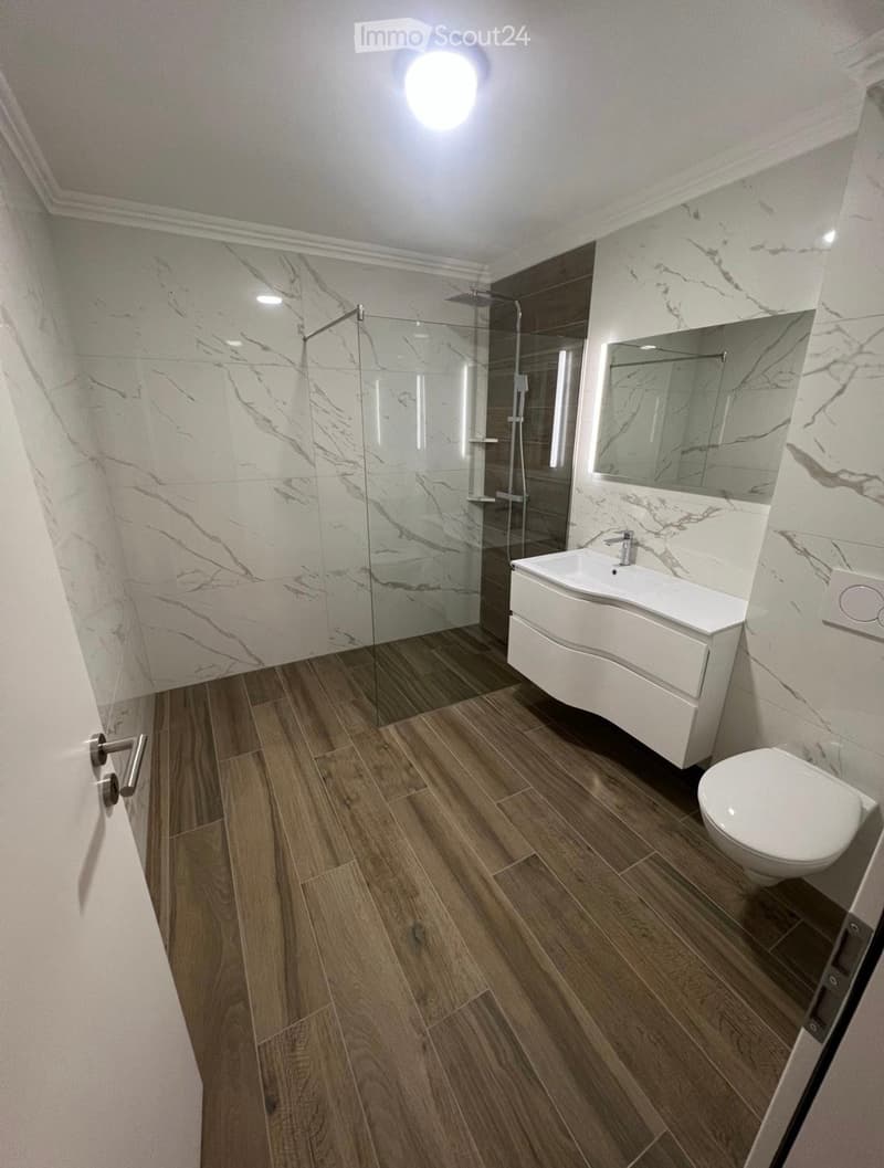 Modern bathroom with marble walls, wooden flooring, a white sink, toilet, and a glass-enclosed shower area.