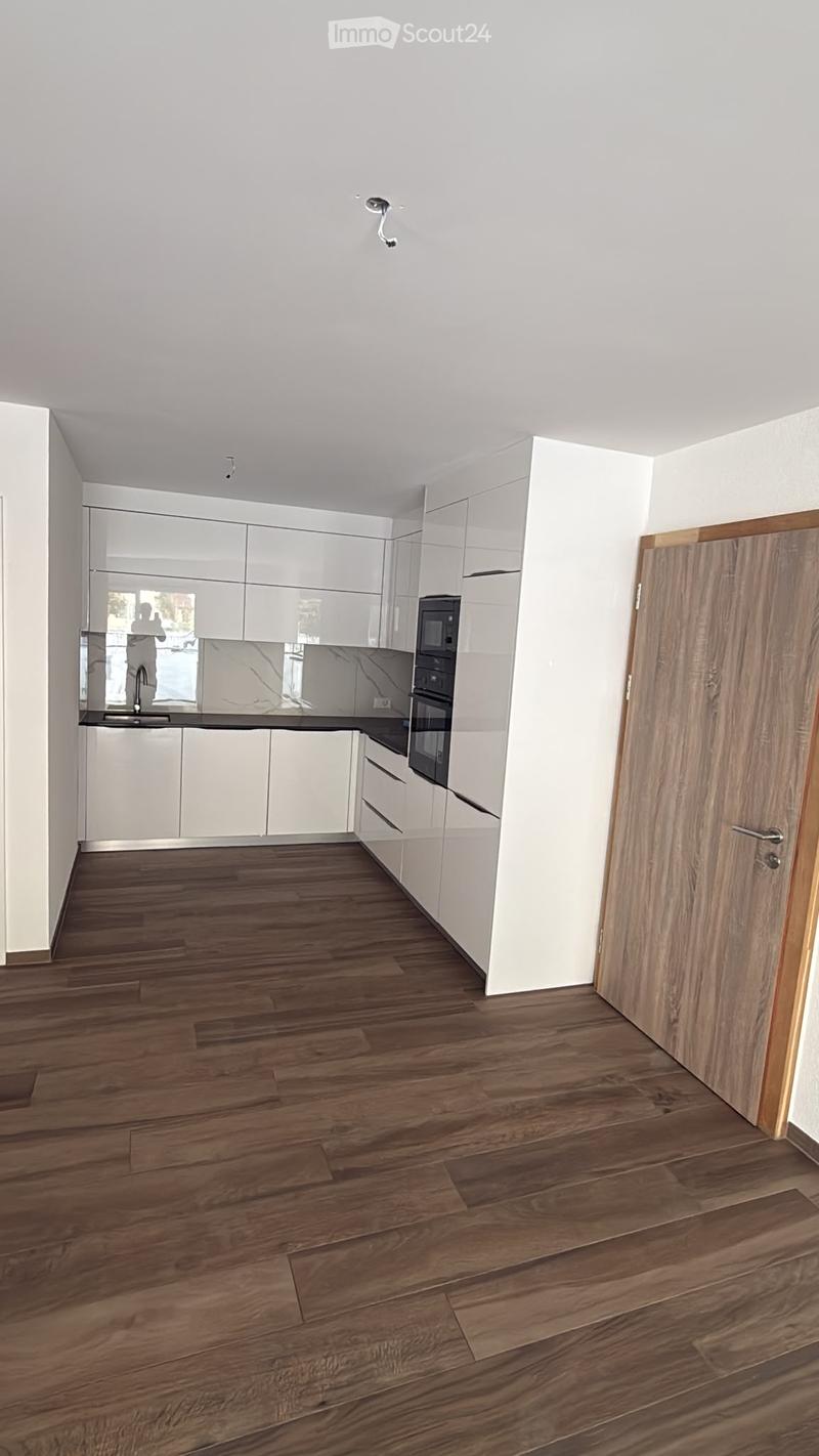 Modern, minimalist kitchen with white cabinets, a dark countertop, and a wooden floor. The space is empty, with no appliances or furniture visible.