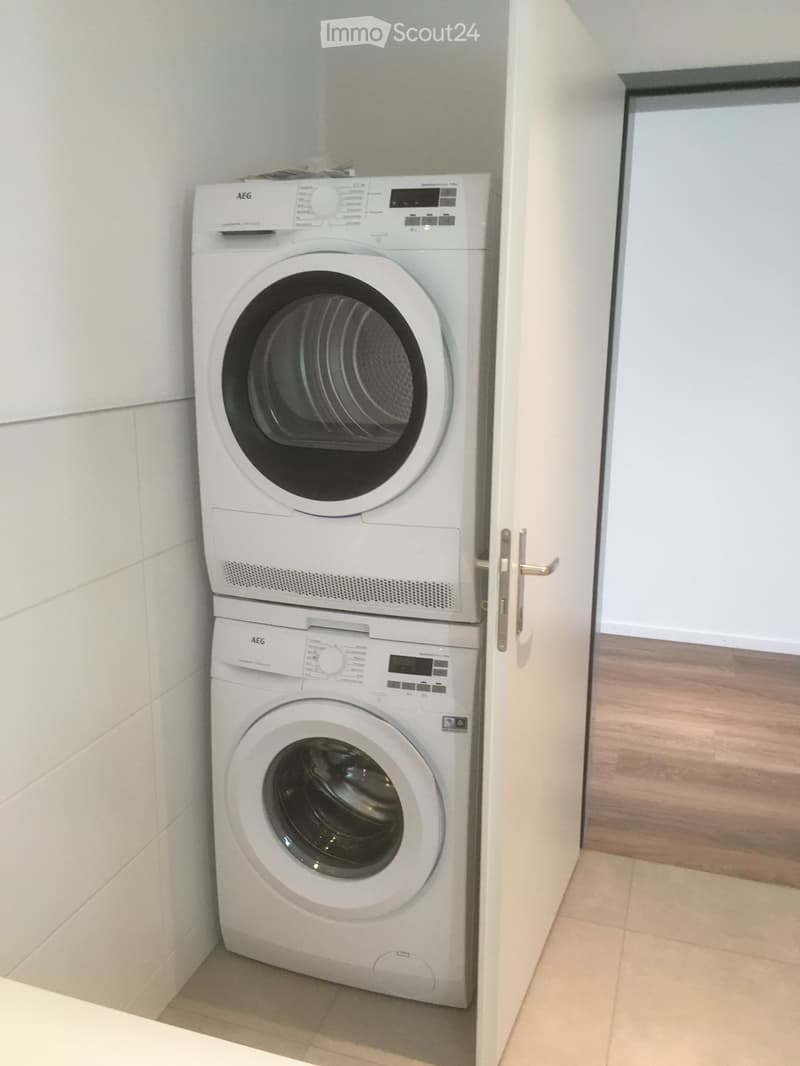A compact bathroom with a stacked washing machine and dryer unit, white tiled walls, and wooden flooring.