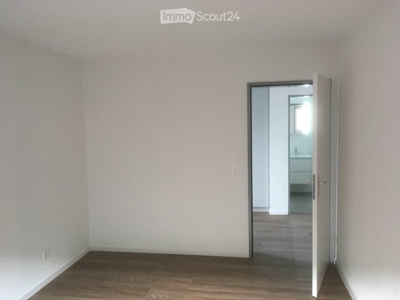 Empty room with wooden flooring and white walls, featuring an open door leading to a small bathroom.
