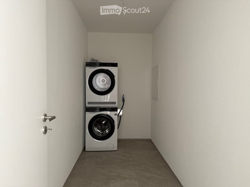 A small, clean room with a washing machine and dryer stacked vertically, light-colored walls, and a tiled floor.