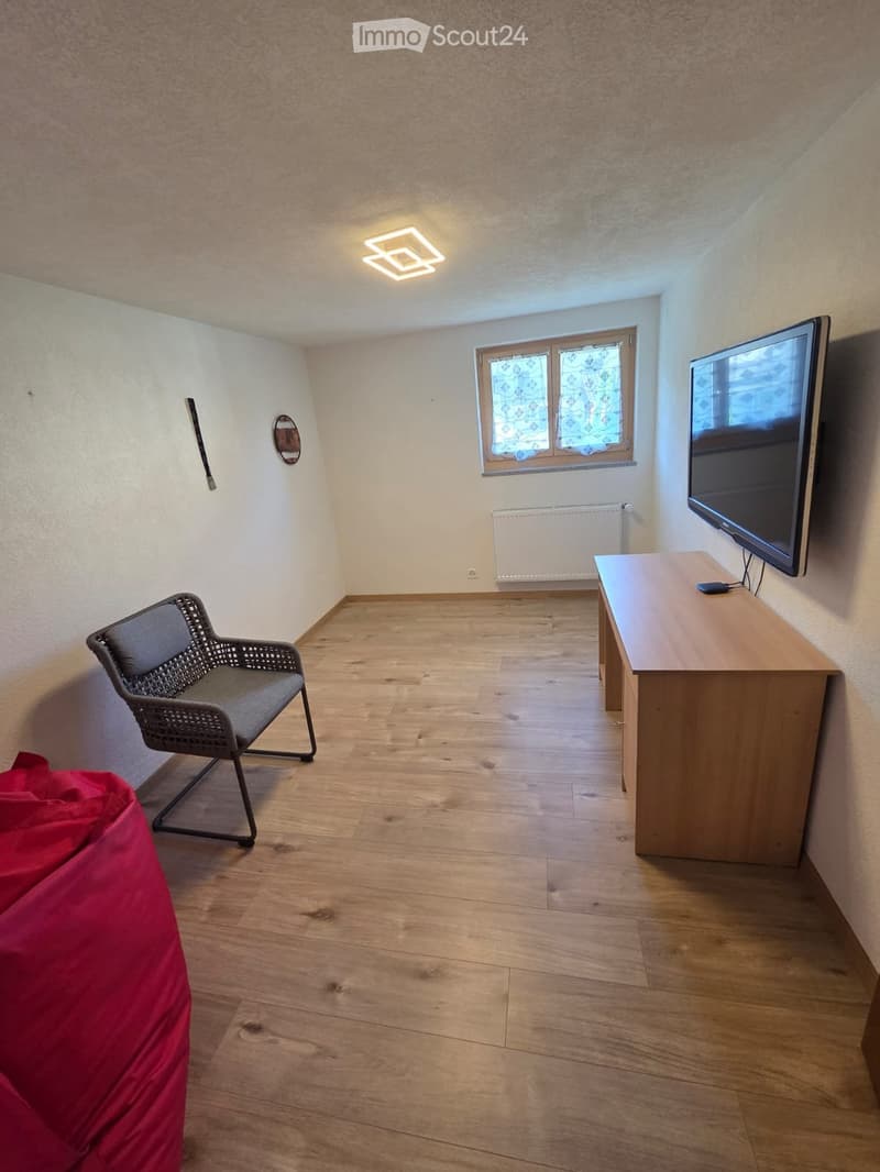 A small, furnished room with wooden flooring, a wicker chair, a wooden table with a TV on top, and a red bean bag chair.