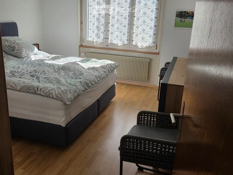 Cozy bedroom with a double bed, patterned duvet, wooden floor, and a wicker chair near a window with patterned curtains.