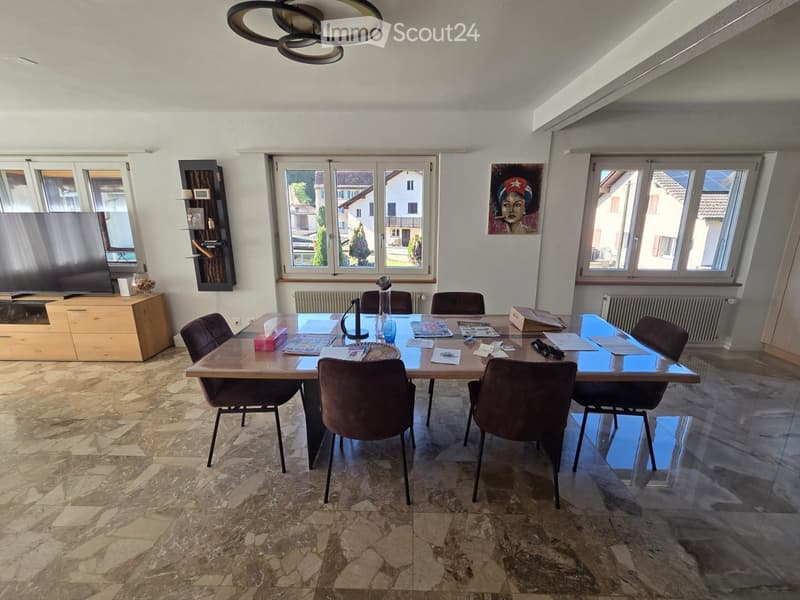 Bright room with large windows, marble floor, a long wooden table surrounded by chairs, a TV on a wooden stand, and a shelf with decorative items.
