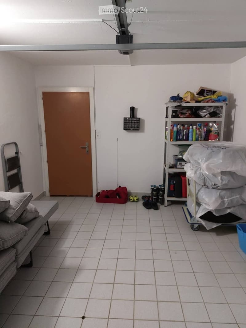 A small, furnished room with tiled flooring, a sofa on the left, a shelf with cleaning supplies, a door, and a cart with bags and shoes.