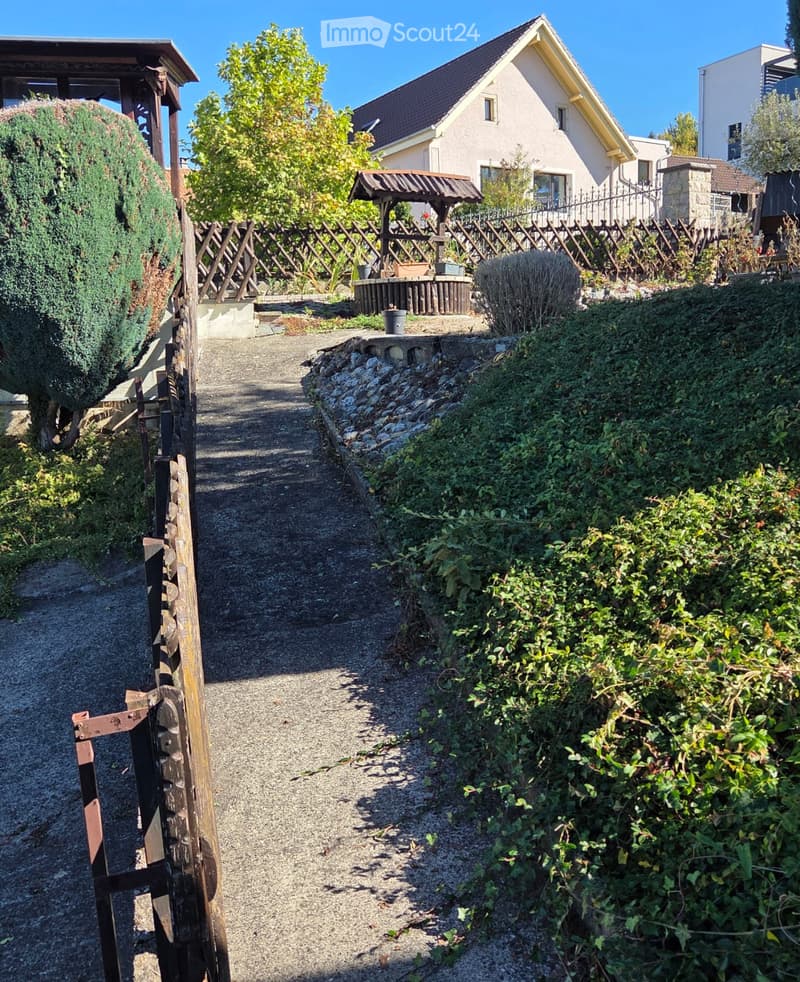 A gravel pathway leads to a wooden gazebo surrounded by greenery and shrubs, with a house visible in the background.