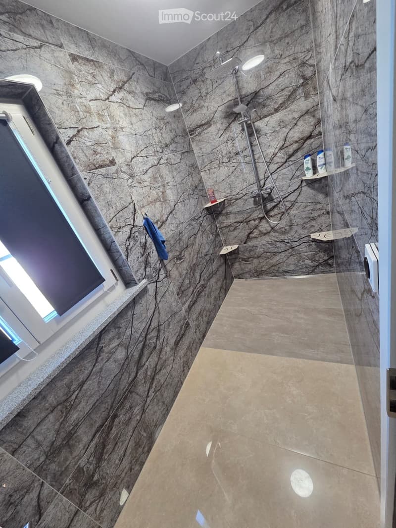 Modern bathroom with marble-patterned walls, a glass-enclosed shower area, a built-in shelf holding toiletries, and a towel hanging on the wall.