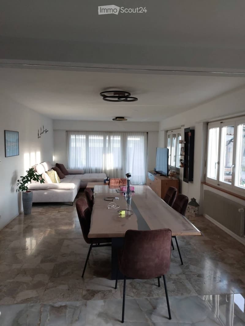 Bright living and dining area with a white sofa, wooden dining table, chairs, TV, and large windows with curtains.