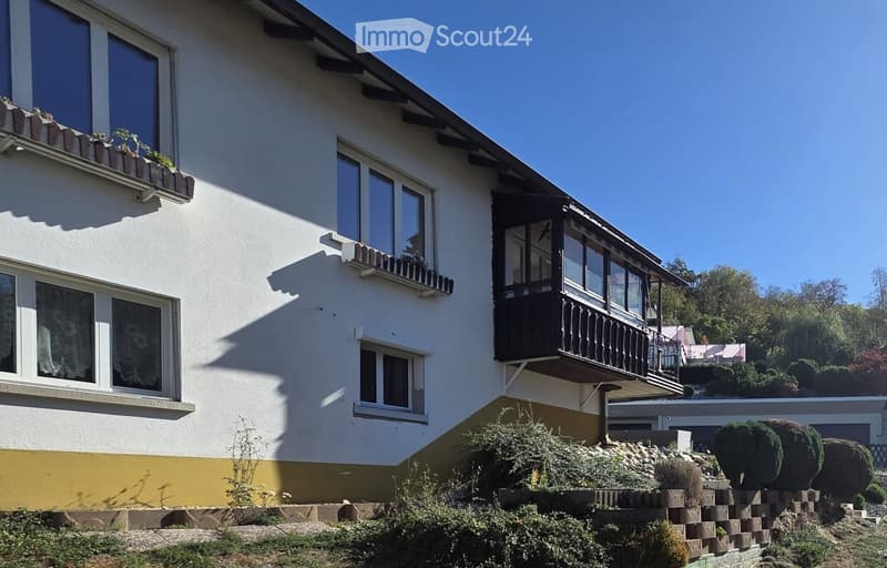 Two-story house with white walls, multiple windows, and a balcony with black railing. The house has a yellow lower section and is surrounded by greenery and a stone wall.