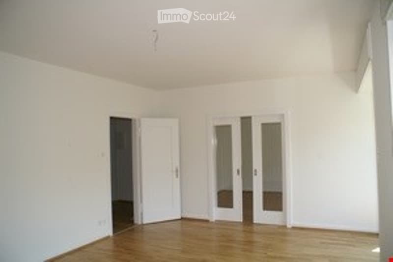 Empty room with white walls, wooden flooring, and a large window with glass doors leading to a balcony.