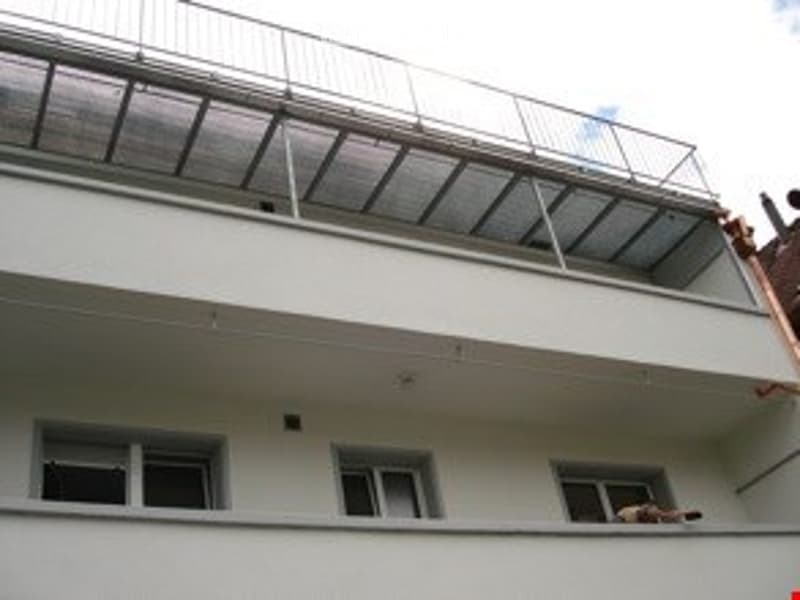 Balcony with glass panels and metal railing, attached to a white building with multiple windows.