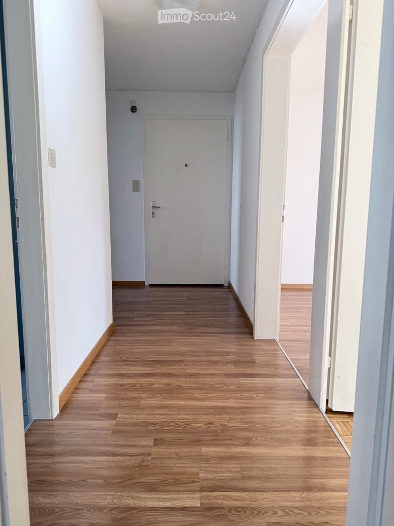Clean, well-lit hallway with light wooden flooring and white walls. A closed door is visible at the end of the corridor.