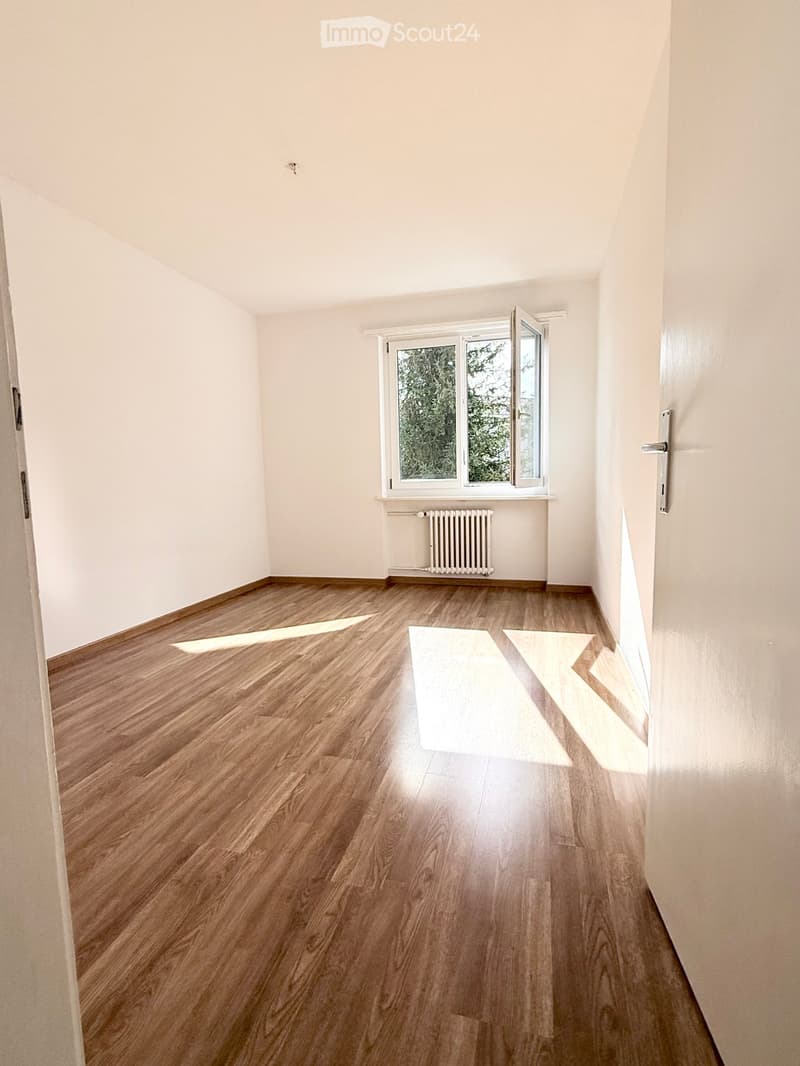 Empty room with light wooden flooring, white walls, and a window with a radiator below it.